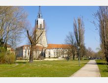 Hauptgebäude mit markantem Turm und gläsernem Aufbau, umgeben von einer parkähnlichen Grünfläche mit Bäumen, Rasenflächen und einem Gehweg. Der sonnige Himmel zeigt eine klare, offene Atmosphäre.