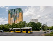 Straßenszene mit einer gelben Straßenbahn im Vordergrund, die eine Kreuzung passiert. Im Hintergrund steht ein markantes Hochhaus mit gelber Fassade und farbigen Fensterelementen, umgeben von Bäumen und weiteren Gebäuden.