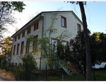 Zweistöckiges Wohnhaus mit hellbeiger Fassade und rot umrahmten Fenstern, umgeben von Bäumen und Büschen. Im Vordergrund verläuft ein niedriger Zaun entlang eines Gehwegs, und im Garten sind Treppen sowie Gartenmöbel sichtbar.