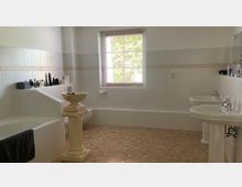 Helles Badezimmer mit beiger Bodenfliese und weißen Wandfliesen, ausgestattet mit einer Badewanne, einem Doppelwaschbecken, einem kleinen Fenster mit weißem Rahmen und einer dekorativen Steinsäule mit Schale. Auf der Ablagefläche befinden sich Pflegeprodukte.