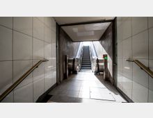 Unterirdischer Durchgang mit weißen Fliesenwänden, Handläufen an den Seiten und einer zentralen Rolltreppe, die zu einem helleren Bereich führt. Die Umgebung ist schlicht und funktional, mit metallischen Elementen an der Rolltreppe.