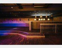 Gemütlicher Veranstaltungssaal mit dunklem Holzboden, einer erhöhten Bühne mit Podest und einer Wand mit geometrischen Spiegelelementen. An der Decke hängen mehrere moderne Lampen, und rechts verläuft eine Metallbrüstung.