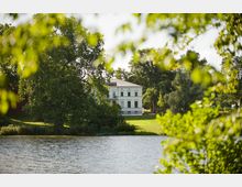 Villa am Seeufer, umgeben von Bäumen und einer Grünfläche. Der Vordergrund zeigt ruhig fließendes Wasser, eingerahmt von unscharfen Blättern.
