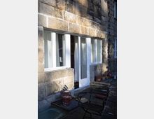 Steinfassade eines Gebäudes mit großen weißen Fenstern und einer offenen Glastür. Im Vordergrund befinden sich eine Terrasse mit Steinplatten, ein Holzstuhl, ein Tisch und mehrere Blumentöpfe.