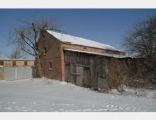 Ländlicher Hofbereich im Winter mit einem roten Backsteingebäude und einem angebauten Holzschuppen, dessen Dach mit Schnee bedeckt ist. Im Vordergrund liegt eine verschneite Fläche mit Fußspuren, umgeben von kahlen Bäumen und Sträuchern unter klarem blauem Himmel.