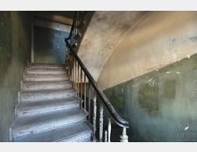 Abgenutztes Treppenhaus mit verwitterten, hellen Holzstufen und einem dunkel lackierten Holzgeländer. Die Wände sind blassgrün gestrichen, mit stellenweise abblätterndem Putz und sichtbaren Alterungsspuren.