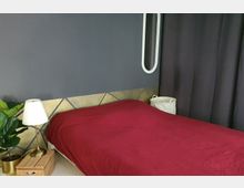 Schlafzimmer mit dunkler Wand, einem Bett mit roter Decke und minimalistischer Dekoration. Links stehen eine Tischlampe auf einer Holzkiste und eine Zimmerpflanze, rechts hängt ein ovaler Spiegel an der Wand.