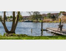 Uferbereich mit einem Holzsteg, auf dem eine Schubkarre steht, im Vordergrund ein Baum und Wiese. Im Hintergrund ein Fluss mit einem Boot sowie Häuser und Schilf am gegenüberliegenden Ufer.