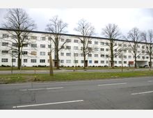 Straßenansicht eines vierstöckigen weißen Wohngebäudes mit flachem Dach, regelmäßig angeordneten Fenstern und mehreren Hauseingängen. Im Vordergrund sind eine Straße, Fußwege, kahler Baumbestand und Grünflächen zu sehen.