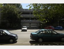 Ein mehrstöckiges Parkhaus mit offenen Seiten, umgeben von Bäumen und in einer städtischen Umgebung. Im Vordergrund stehen geparkte Autos auf einer Straße, während die Einfahrt des Parkhauses mit Schranken und Verkehrsschildern zu sehen ist.