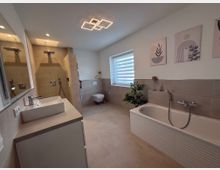 Modernes Badezimmer mit beige gefliesten Wänden und Böden. Das Zimmer beinhaltet eine Dusche mit Regenduschkopf, ein weißes Waschbecken auf einem Schrank, eine Badewanne mit integrierter Armatur und ein hängendes WC; an der Wand sind minimalistische Bilder und ein Fenster mit Lamellenrollo zu sehen.