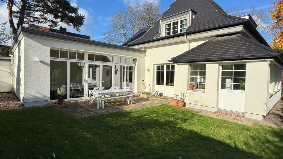 Rückansicht eines hell verputzten Einfamilienhauses mit dunklem Walmdach und großen Sprossenfenstern, teilweise als verglaster Anbau ausgeführt. Davor liegt eine gepflasterte Terrasse am Rasen, umgeben von Bäumen unter blauem Himmel.