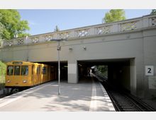 Eine hellgelbe U-Bahn hält an einem offenen, teilweise überdachten Bahnsteig mit der Beschilderung "2" an der Wand. Über dem Bahnsteig verläuft eine helle Betonbrücke mit dekorativen Geländern, umgeben von grünen Bäumen und klarem Himmel.