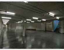 Großer unterirdischer Raum mit Betonwänden und -decke, beleuchtet durch Reihen von Leuchtstofflampen. Der Boden ist dunkel und glänzend, es gibt mehrere Türen, eine Sitzbank und eine Treppe, die nach oben führt.