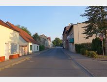 Straßenszene in einem Dorf oder Vorort mit niedrigen Wohn- und Geschäftshäusern auf beiden Seiten, teils mit roten Ziegeldächern. Der asphaltierte Weg ist von Gehwegen, Grünflächen und einzelnen Bäumen gesäumt, die bei Tageslicht sichtbar sind.