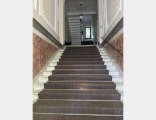 Innenansicht eines historischen Treppenhauses mit breiter Marmortreppe und dunklem Teppichläufer, flankiert von marmorverkleideten Wänden und Handläufen. Oben sind Stuckdecken und hängende Kugellampen zu sehen, am Ende des Flurs ein großes Fenster mit farbigem Glas.