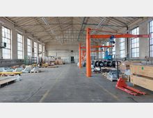 Eine geräumige Industriehalle mit hohen Decken und großen Fenstern an den Seitenwänden. Im Vordergrund befinden sich Paletten mit Materialien, Kisten und ein roter Hubwagen; auf der rechten Seite sind orangefarbene Laufkrane zu sehen, die Metallrollen und andere Gegenstände tragen.