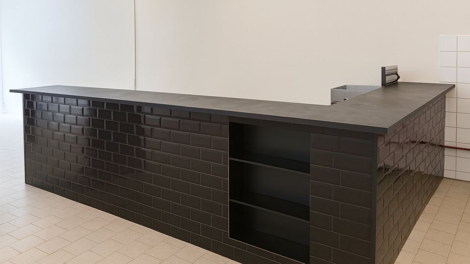 Moderne Küchen- oder Barzeile in L-Form mit schwarzer Arbeitsplatte und Fronten aus glänzenden, schwarzen Rechteckfliesen, inklusive offenem Regalfach. Heller Raum mit beigem Fliesenboden und weißen Wänden, im Hintergrund teils weiß geflieste Wandbereiche.