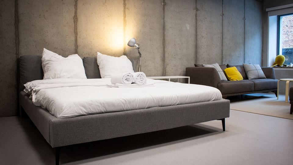 Modernes Schlafzimmer im Loft-Stil mit Betonwänden, großem grauen Polsterbett mit weißer Bettwäsche und zusammengerollten Handtüchern sowie einer Stehlampe am Nachttisch. Im Hintergrund stehen ein graues Sofa mit Kissen und ein großes Fenster, das Tageslicht in den Raum lässt.