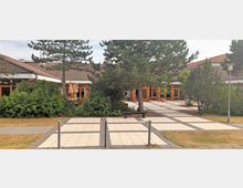 Hofbereich eines einstöckigen Gebäudes mit hellbraunen Dachziegeln und großen, holzgerahmten Fenstern. Im Vordergrund ist ein gepflasterter Platz mit Bäumen, Sträuchern und Sitzbänken, umgeben von Laternen und Gehwegen.