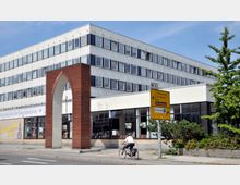 Modernes Bürogebäude mit weißer Fassade und zahlreichen quadratischen Fenstern; im Vordergrund ein roter, spitzbogiger Backsteinbogen sowie ein Verkehrsschild mit Wegweisern zu lokalen Zielen. Ein Fahrradfahrer fährt auf der Straße entlang, umgeben von etwas Grün.