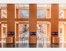 Innenbereich eines modernen Gebäudes mit einer Holz- und Glasarchitektur. Zu sehen sind große vertikale Holzpaneele, eine gläserne Dachkonstruktion mit Metallic-Elementen und Durchgänge mit Drehkreuzen im unteren Bereich.