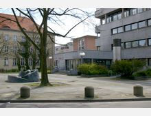 Hofbereich mit Betonpflasterung, einer modernen abstrakten Skulptur und Treppenaufgang zu einem flachen, grau verputzten Gebäude mit großen Fenstern; links im Hintergrund ein historisches Gebäude mit Satteldach und hell verputzter Fassade.