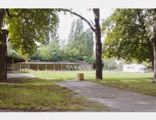 Ein leerer Außenbereich mit einer überwucherten Wiese, vereinzelten Bäumen und einem gepflasterten Weg. Im Hintergrund steht ein langes, flaches Gebäude mit Graffiti, umgeben von dichter Vegetation.