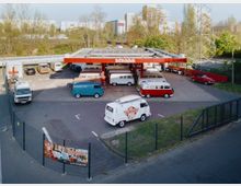 Eine Tankstelle mit rotem Dach, erkennbar als "Bulli-Tankstelle", auf deren Gelände mehrere VW-Busse verschiedener Modelle und Farben geparkt sind. Im Vordergrund ist ein Zaun mit Werbebannern sowie einige Bäume und Gebäude im Hintergrund zu sehen.