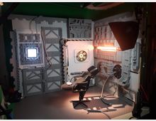 Ein Raum, der wie das Innere eines futuristischen Raumschiffs gestaltet ist, mit grauen Wänden, strukturierten Paneelen, Kabeln, einer kleinen beleuchteten Öffnung, einem beleuchteten Steuerungsmodul und einem schwarzen Drehstuhl in der Mitte. Beleuchtungselemente und technische Details betonen das Hightech-Design.