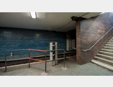 U-Bahn-Station mit einer Wand aus blauen Kacheln, neben der sich ein Treppenaufgang mit roten Backsteinwänden befindet. Im Vordergrund sind Geländer und eine Metallkonstruktion, im Hintergrund ist ein Gleis zu sehen.