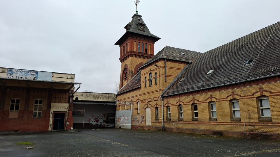 Backsteingebäudekomplex mit langem, ziegelgedecktem Satteldach und Rundbogenfenstern, dahinter ein hoher Turm mit Uhr und spitzem Dach. Davor liegt ein leerer, asphaltierter Hof; an einigen Wänden sind verwitterte Flächen und Graffiti zu sehen.