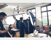 Ein Fotostudio mit mehreren Lichtquellen, Reflektoren und einem schwarzen Hintergrund, eingerichtet in einem Raum mit hohen Fenstern und einem Kronleuchter. Drei Personen arbeiten an der Fotoausrüstung und einem Tisch mit einem Laptop, während weitere Möbel und Dekorationen im Hintergrund sichtbar sind.