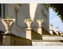 Eine Backsteinfassade mit Fenstern im historischen Stil, davor eine Treppe mit niedrigen Mauern, die mit verzierten Steinvasen mit Pflanzen bestückt sind. Die Szene befindet sich im Freien bei Tageslicht, teilweise im Schatten von Bäumen.