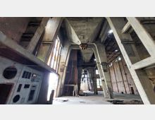 Verlassene Industriehalle mit massiven Betonträgern, hohen Decken und viel sichtbarer Stahl- und Ziegelkonstruktion. Links im Bild sind Metalltafeln mit alten Schaltflächen und Fenstern zu sehen; Licht dringt durch große Fensteröffnungen.