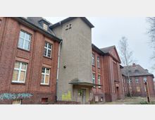 Backstein-Gebäude mit mehreren Etagen, Fenstern, teilweise mit Holzbrettern vernagelt. In der Mitte ein grauer Anbau mit Flachdachvordach, umgeben von kahlen Bäumen.