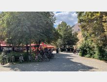 Städtischer Platz mit Kopfsteinpflaster, gesäumt von Bäumen und Fahrrädern. Im Hintergrund befinden sich Cafés mit roten Sonnenschirmen sowie mehrstöckige Wohngebäude, teilweise verdeckt durch das dichte Grün.