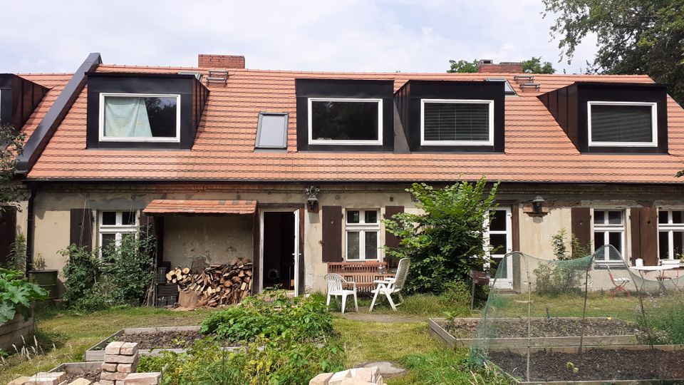 Rückansicht eines niedrigen, länglichen Wohnhauses mit rotem Ziegeldach und mehreren dunklen Gaubenfenstern sowie Holzläden an den Fenstern. Davor liegt ein Garten mit Hochbeeten, Rasen, gestapelten Ziegeln und einem kleinen Sitzbereich neben einem überdachten Holzlager.