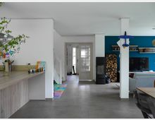 Moderne Wohnküche mit offenem Grundriss, weiß gestrichenen Wänden und einer blauen Akzentwand im Wohnzimmerbereich. Im Vordergrund befindet sich eine helle Küchenzeile, rechts ein Essbereich mit Holzstuhl, im Hintergrund ein Kaminofen, gestapeltes Feuerholz und ein Wohnzimmer mit Sofa und Fernseher.