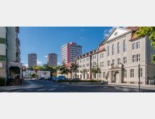 Straßenszene mit einem großen historischen Gebäude rechts, beschriftet als "Gemeindehaus von St. Georg", mit einer weißen Fassade und roten Dächern. Im Hintergrund sind modernere Hochhäuser sichtbar, während die Straße von geparkten Autos gesäumt ist, umgeben von Bäumen und kleineren Gebäuden.