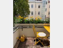 Kleiner Balkon mit Holzfliesen, gelbem Klapptisch und -stuhl sowie einer gelben Gießkanne; entlang des Geländers stehen Blumenkästen mit bunten Blumen. Im Hintergrund sind ein großer Baum und die Fassade eines gegenüberliegenden Wohngebäudes mit mehreren Fenstern zu sehen.