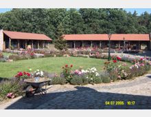 Ein Innenhof mit einem gepflegten Blumengarten, umgeben von einer hölzernen Stallanlage mit roten Ziegeldächern. Im Vordergrund stehen eine Schubkarre und gepflasterte Wege, im Hintergrund befindet sich ein Wald.