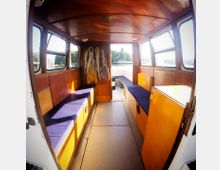 Innenraum eines kleinen Bootes mit Holzwänden und -decken, seitlichen Fenstern und blauen Sitzpolstern. An einer Wand hängen mehrere zusammengerollte Seile; durch die Fenster ist Wasser und eine grüne Uferlandschaft sichtbar.