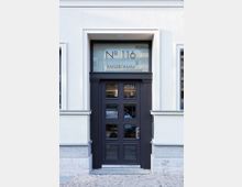 Eingangsbereich eines Gebäudes mit weißer Fassade, dunkler Tür mit quadratischen Glaseinsätzen und einer Oberlichtscheibe, auf der "No 116 Kaiserdamm" steht. Pflastersteine säumen den Bereich vor der Tür.