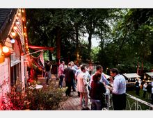 Eine Außenveranstaltung in einem bewaldeten Garten mit Lichterketten und mehreren Menschen, die sich auf einer Holzterrasse unterhalten. Im Hintergrund sind Marktstände und weitere Besucher sichtbar.