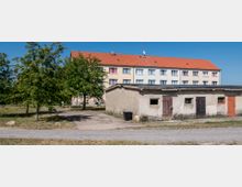 Einfache Wohnsiedlung mit einem hellen Mehrfamilienhaus im Hintergrund, gelbem Putz und rotem Ziegeldach. Im Vordergrund steht eine kleine, flache Betonstruktur mit alten Holztüren und Fenstern, umgeben von Bäumen, Grasflächen und einem Schotterweg.