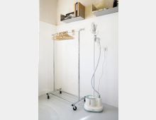 Eine minimalistische Ecke in einem Raum mit einem mobilen Kleiderständer aus Metall, an dem mehrere Holzkleiderbügel hängen, und einem weißen Dampfbügler auf dem Boden. Darüber befinden sich an der Wand zwei schlichte Regalböden mit Büchern und Dekoration.