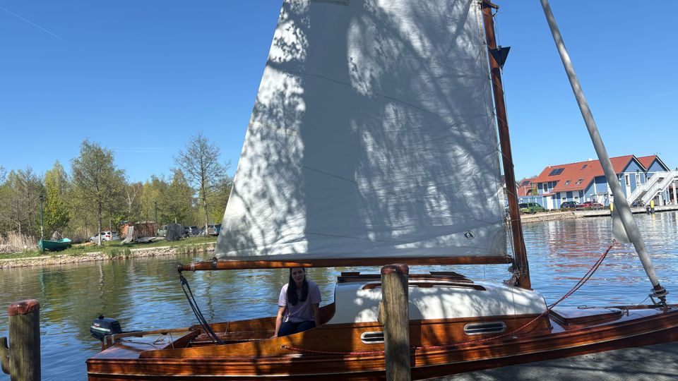 Holz-Segelboot mit großem weißem Segel liegt an einem Steg in einem ruhigen Hafenbecken. Im Hintergrund stehen Bäume am Ufer sowie mehrere blau-weiße Häuser mit roten Dächern unter klarem Himmel.