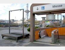 U-Bahnhof-Eingang mit Rolltreppe und orange gekachelten Wänden, darüber ein Schild mit der Aufschrift „Messe“. Im Hintergrund befinden sich ein moderner Ausstellungsbau, ein Freigelände mit Sitzbänken und der Funkturm.