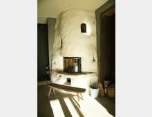 Ein rustikaler Raum mit einem weiß verputzten, abgerundeten Kachelofen als Hauptmotiv. Der Ofen hat ein kleines Fenster und eine gewölbte Form, daneben stehen ein Holzschemel, ein Besen und ein Eimer auf einem Betonboden.